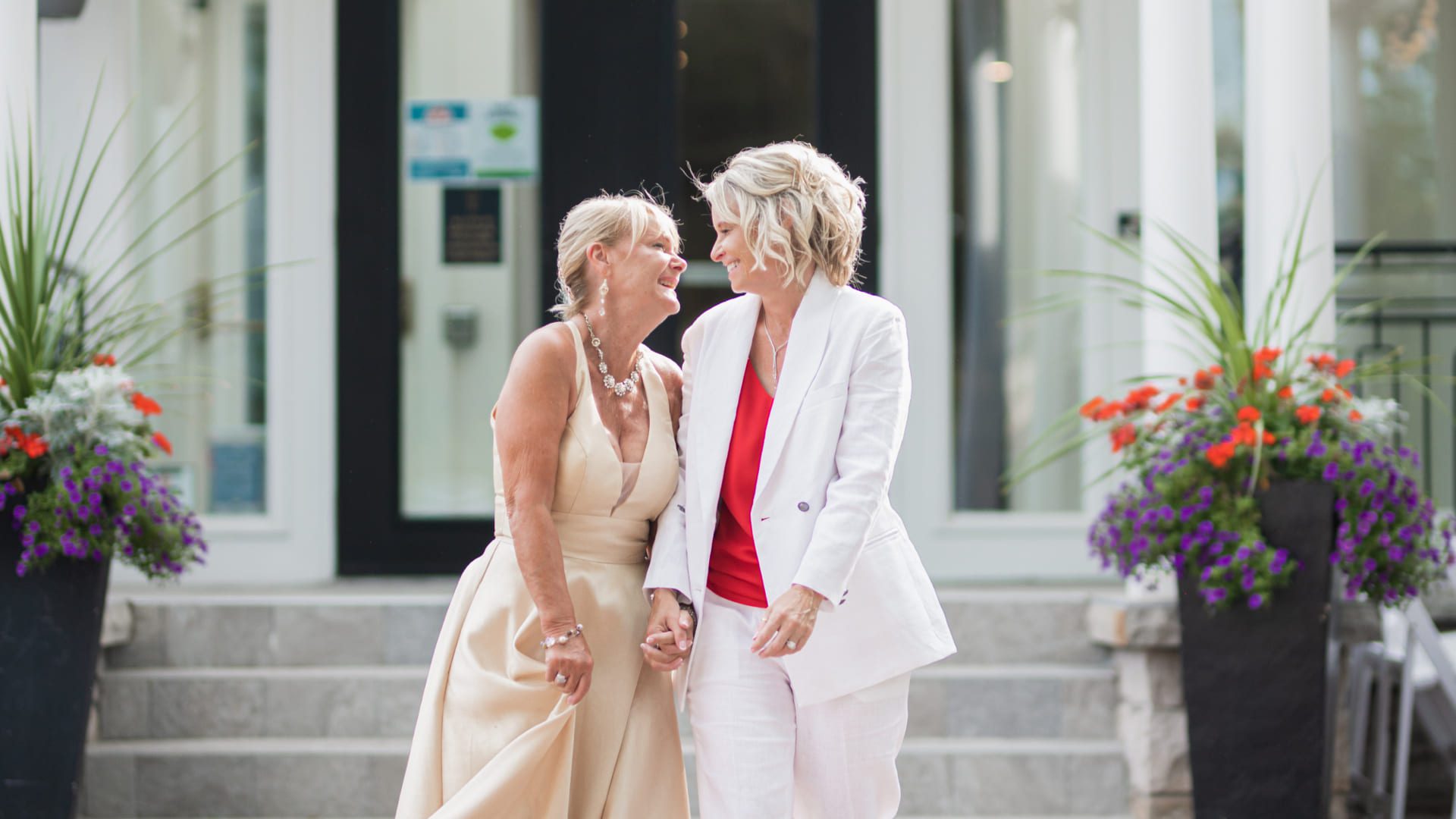 Two women smiling at each other while standing close together. One is in a beige dress, and the other in a white suit with a red top. They are on a stone pathway flanked by tall planters with flowers, in front of a building with large doors. 124 on Queen Hotel and Spa | Hotel & Spa Niagara-on-the-lake | 1 (855) 988-4552