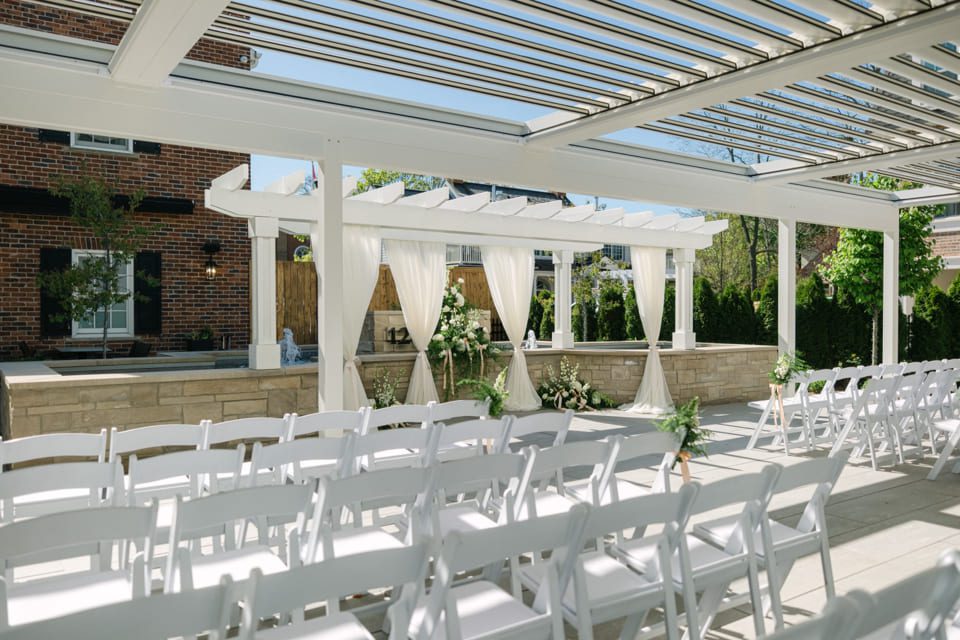 Outdoor wedding venue with rows of white chairs facing a decorated arbor with white drapes and floral arrangements. Sunny, clear skies enhance the garden-like setting with a brick building in the background. 124 on Queen Hotel and Spa | Hotel & Spa Niagara-on-the-lake | 1 (855) 988-4552