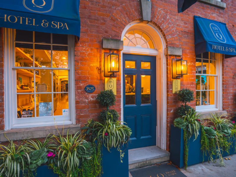 Quaint brick building with Hotel & Spa signage. Features blue awnings, a dark blue door, and warm lit lanterns. Lush planters with various greenery adorn the entrance. Windows display cozy interior details. House number 125 visible. 124 on Queen Hotel and Spa | Hotel & Spa Niagara-on-the-lake | 1 (855) 988-4552