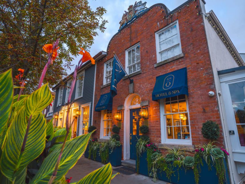 A charming hotel and spa in a historic brick building, featuring blue awnings and elegant signage. The facade is adorned with lush greenery and colorful flowers. Nearby trees add to the inviting, quaint atmosphere. 124 on Queen Hotel and Spa | Hotel & Spa Niagara-on-the-lake | 1 (855) 988-4552
