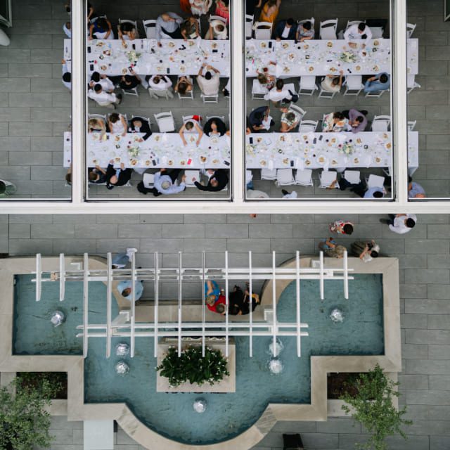 Aerial view of two long tables with people seated, having a meal. The tables are set with white tablecloths and dishes. A decorative water feature with plants and fountains is in the foreground. The scene is outdoors on a paved area. 124 on Queen Hotel and Spa | Hotel & Spa Niagara-on-the-lake | 1 (855) 988-4552