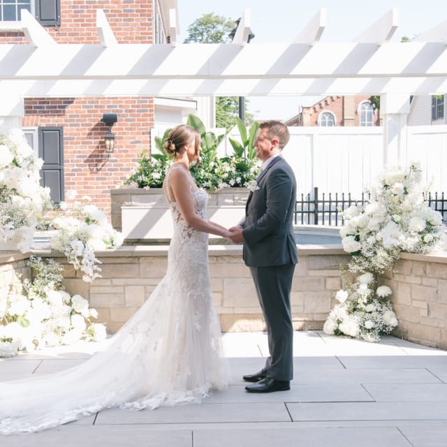 A bride and groom stand facing each other under a sunlit pergola. The bride wears a long, lace wedding dress, and the groom is in a dark suit. They are surrounded by white floral arrangements, and a brick building is visible in the background. 124 on Queen Hotel and Spa | Hotel & Spa Niagara-on-the-lake | 1 (855) 988-4552