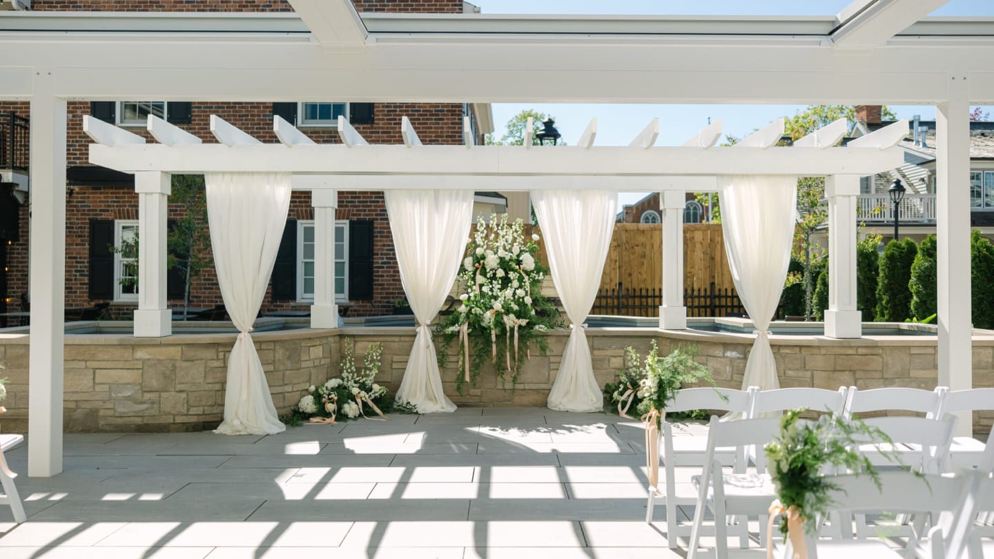An outdoor wedding setup features a white pergola draped with sheer white curtains. Floral arrangements with white and green flowers adorn the area. White chairs with green decorations are neatly arranged in rows, and buildings are visible in the background. 124 on Queen Hotel and Spa | Hotel & Spa Niagara-on-the-lake | 1 (855) 988-4552