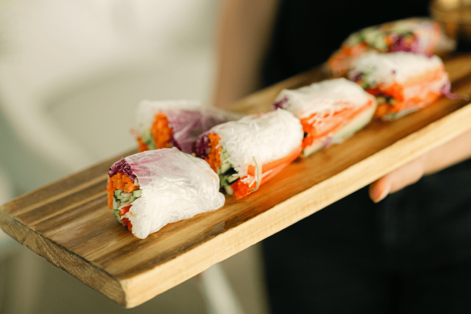 A person holds a wooden tray with four fresh Vietnamese spring rolls. The rolls are filled with rice noodles, carrots, and other vegetables, wrapped in translucent rice paper. The background is softly blurred. 124 on Queen Hotel and Spa | Hotel & Spa Niagara-on-the-lake | 1 (855) 988-4552