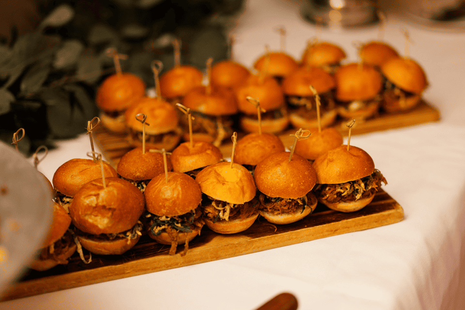 Mini pulled pork sliders are arranged on wooden boards. Each slider is secured with a small skewer, featuring fluffy buns and a visible layer of pulled pork. The boards are placed on a white tablecloth, with some greenery blurred in the background. 124 on Queen Hotel and Spa | Hotel & Spa Niagara-on-the-lake | 1 (855) 988-4552