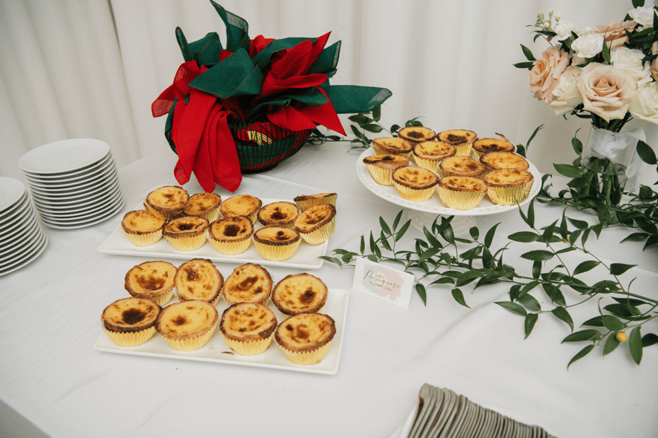 A table displays rows of custard tarts on rectangular and round plates. Nearby are stacked plates, a basket with red and green cloth, leafy greenery, and a bouquet of pale roses. A small card is placed near the tarts. 124 on Queen Hotel and Spa | Hotel & Spa Niagara-on-the-lake | 1 (855) 988-4552