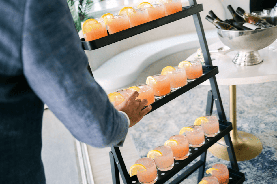 A person reaches for a glass from a multi-tiered stand holding several pink cocktails garnished with orange slices. Nearby, a table has bottles of wine chilling in a metallic bowl. The setting appears casual and elegant. 124 on Queen Hotel and Spa | Hotel & Spa Niagara-on-the-lake | 1 (855) 988-4552