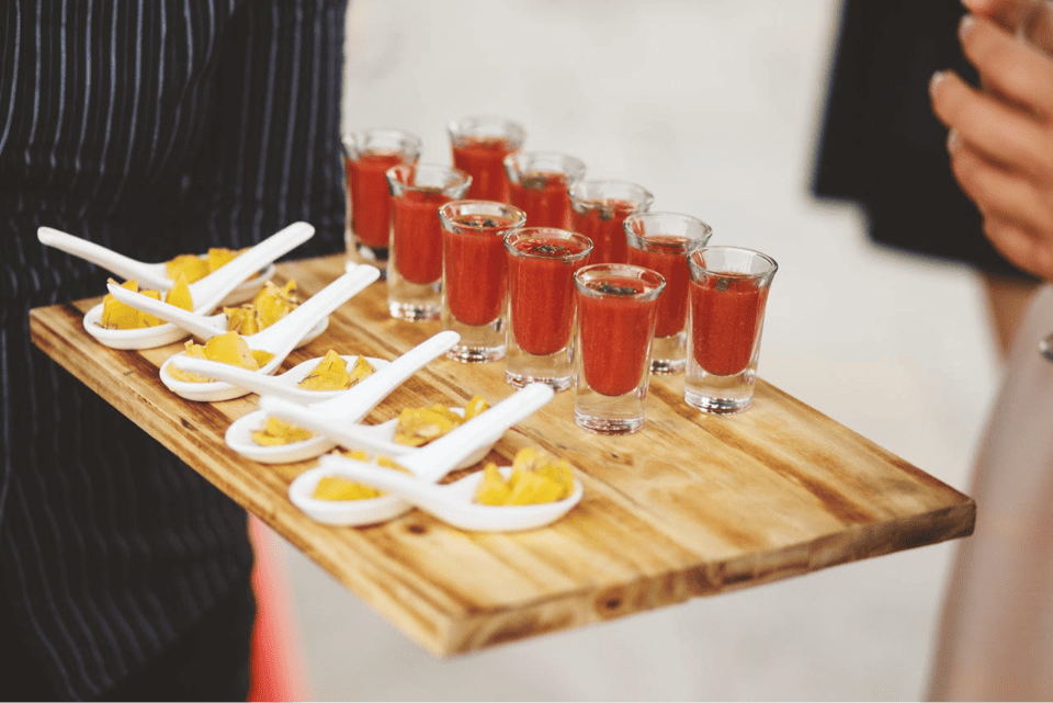 A person holds a wooden tray with neatly arranged small glasses of red liquid, possibly a cocktail or juice, and spoons of food garnished with chopped yellow pieces, possibly mango or cheese. The background is partially blurred. 124 on Queen Hotel and Spa | Hotel & Spa Niagara-on-the-lake | 1 (855) 988-4552