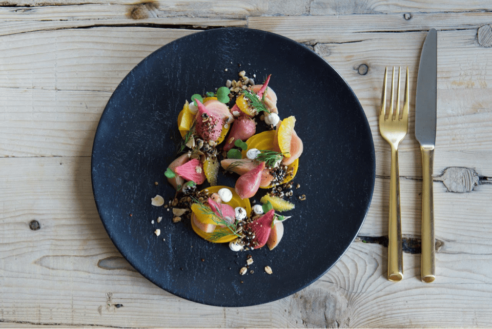 A black plate with a colorful dish featuring sliced beets, orange segments, and garnished with herbs and crumbled nuts. A gold knife and fork are placed on the wooden table beside the plate. 124 on Queen Hotel and Spa | Hotel & Spa Niagara-on-the-lake | 1 (855) 988-4552