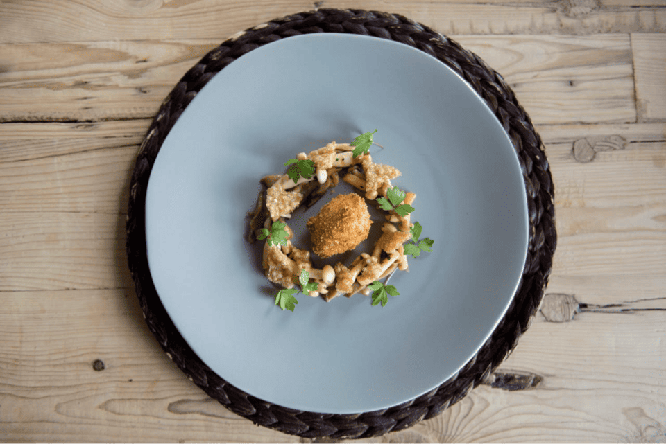 A gourmet dish with a ring of assorted mushrooms and herbs surrounding a central croquette, presented on a sleek blue plate over a woven placemat on a wooden table. 124 on Queen Hotel and Spa | Hotel & Spa Niagara-on-the-lake | 1 (855) 988-4552