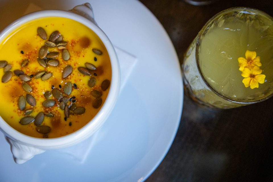 A bowl of creamy orange soup garnished with pumpkin seeds and spices sits on a white plate. Next to it, a glass of light green drink features a small yellow flower as decoration. The surfaces are placed on a dark table. 124 on Queen Hotel and Spa | Hotel & Spa Niagara-on-the-lake | 1 (855) 988-4552