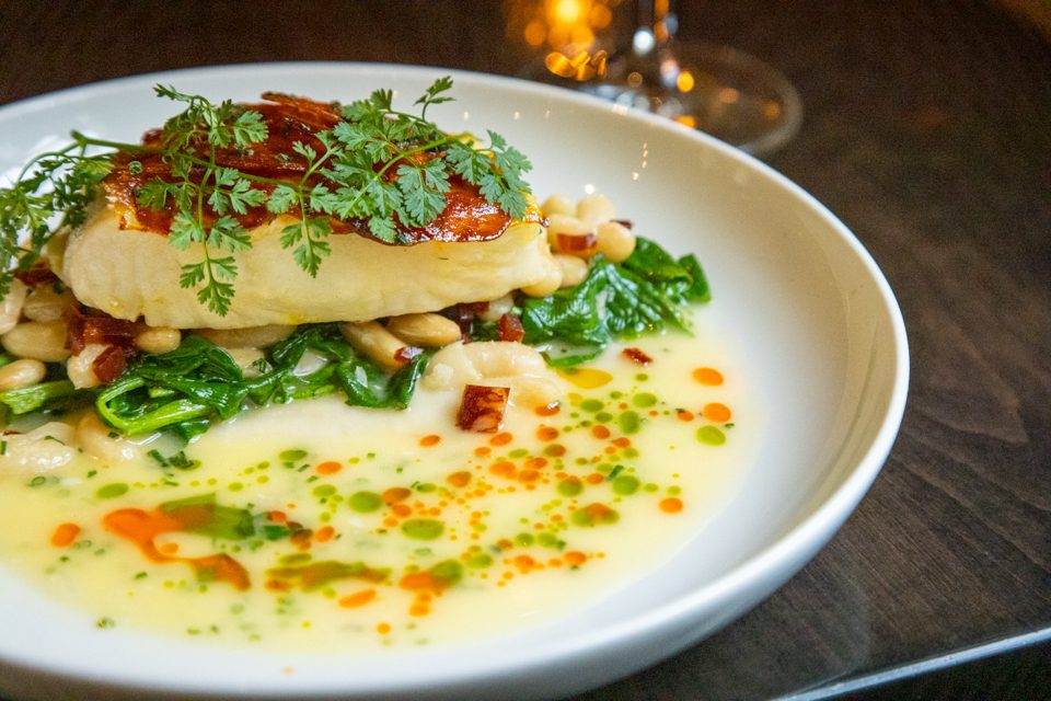 A beautifully plated dish featuring a seared piece of fish topped with fresh herbs, lying on a bed of sautéed spinach and white beans. The plate is garnished with colorful oil droplets and a rich sauce. 124 on Queen Hotel and Spa | Hotel & Spa Niagara-on-the-lake | 1 (855) 988-4552