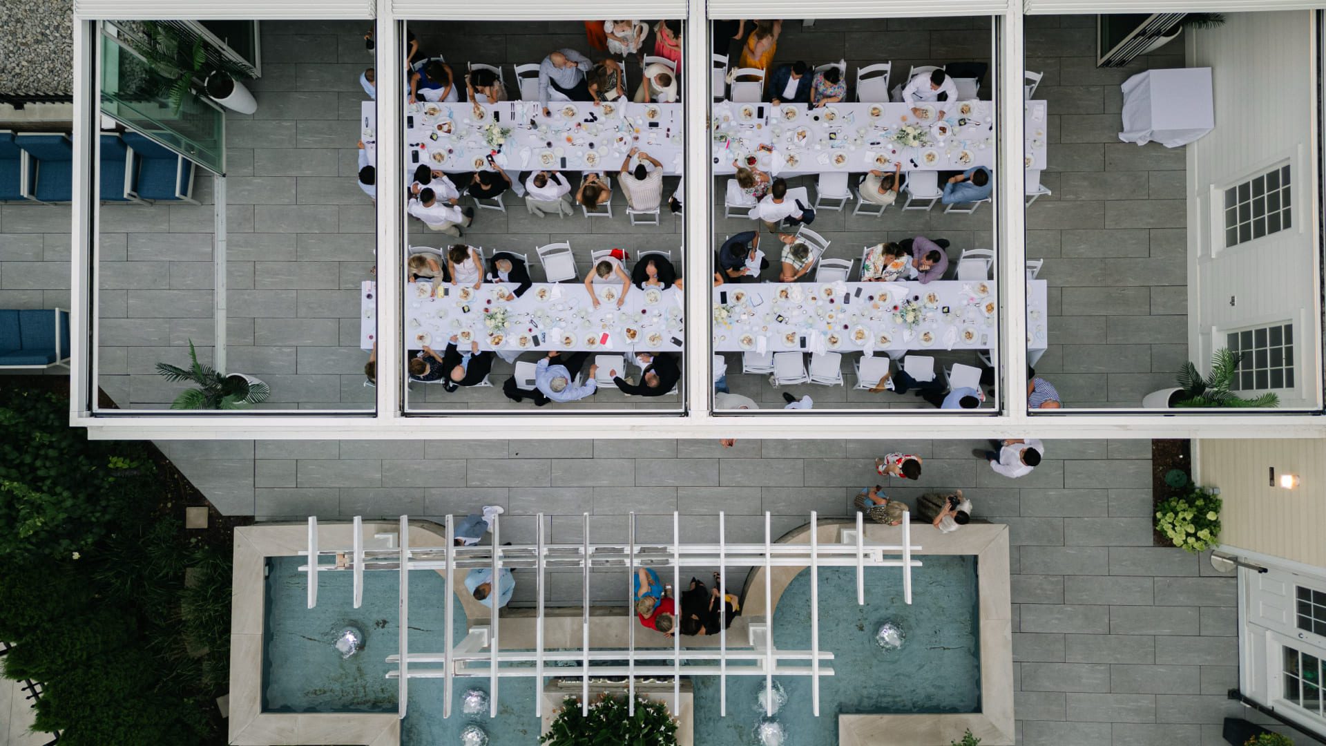 Overhead view of a long dining table with guests seated on a patio. The table is covered with white tablecloths and dishes. Adjacent, there is a decorative water feature with fountains. Lush greenery surrounds the area. 124 on Queen Hotel and Spa | Hotel & Spa Niagara-on-the-lake | 1 (855) 988-4552