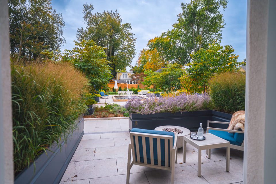 View from a patio with a table and two chairs, overlooking a garden with lush greenery and trees. The background features a distant house and a cloudy sky, creating a serene outdoor setting. 124 on Queen Hotel and Spa | Hotel & Spa Niagara-on-the-lake | 1 (855) 988-4552