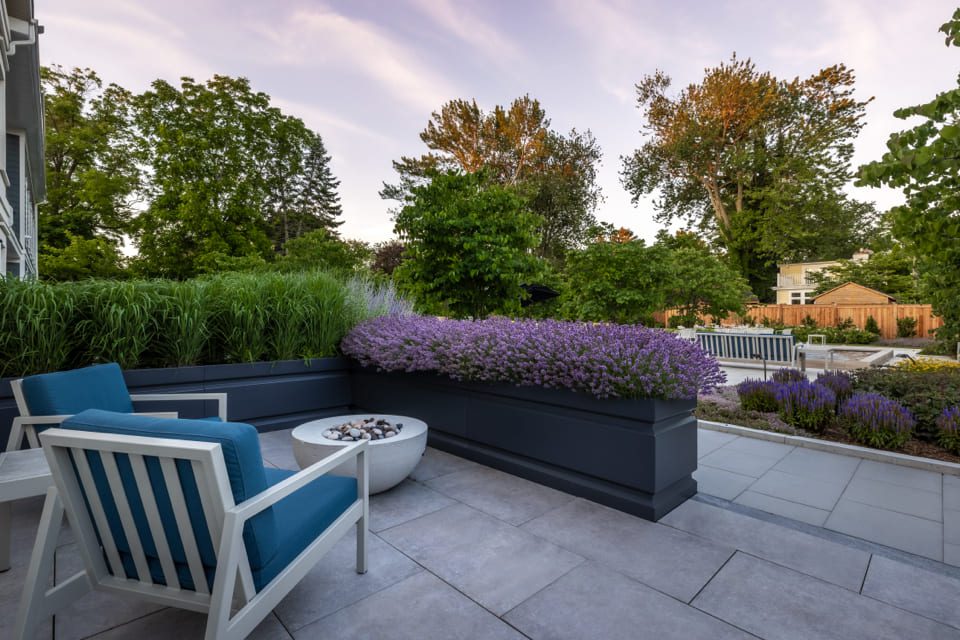 A modern outdoor patio features white and blue chairs facing a round fire pit. Lush green plants and lavender flowers line the patio, offering a serene view of a landscaped garden with tall trees under a clear sky. 124 on Queen Hotel and Spa | Hotel & Spa Niagara-on-the-lake | 1 (855) 988-4552