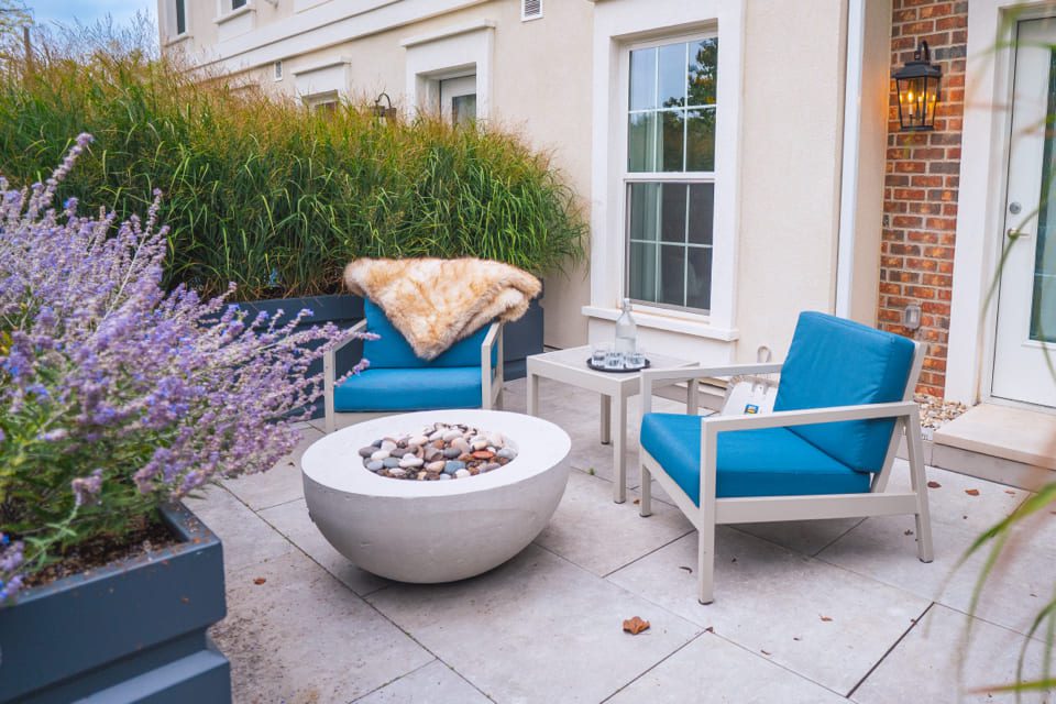 Cozy patio scene with two blue-cushioned chairs, one draped with a fur throw, surrounding a round fire pit filled with stones. A small table holds a lantern and candle. Lush greenery and purple flowers in planters enhance the tranquil setting. 124 on Queen Hotel and Spa | Hotel & Spa Niagara-on-the-lake | 1 (855) 988-4552