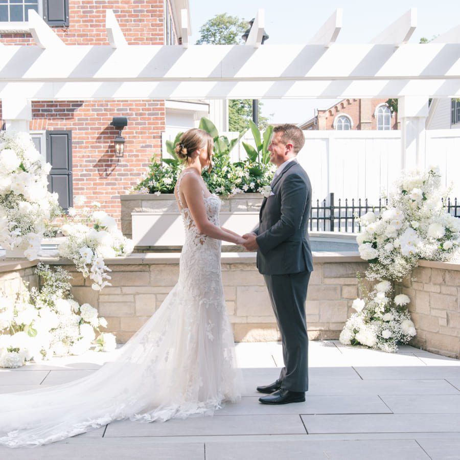 A bride in a white gown and a groom in a dark suit stand facing each other, holding hands, outdoors under a white pergola, surrounded by white floral arrangements. 124 on Queen Hotel and Spa | Hotel & Spa Niagara-on-the-lake | 1 (855) 988-4552