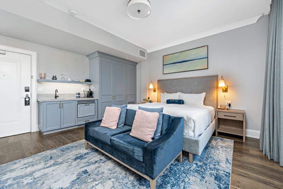 A modern hotel room with a bed, two nightstands, a blue velvet sofa with pink pillows, a blue patterned rug, kitchenette, and soft gray and blue decor. Large windows with curtains let in natural light. 124 on Queen Hotel and Spa | Hotel & Spa Niagara-on-the-lake | 1 (855) 988-4552