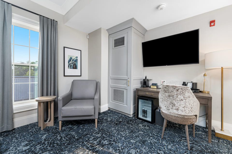 A modern hotel room corner with a gray armchair, small round side table, framed wall art, desk with a chair, mini fridge, coffee maker, and a large flat-screen TV mounted on the wall. 124 on Queen Hotel and Spa | Hotel & Spa Niagara-on-the-lake | 1 (855) 988-4552