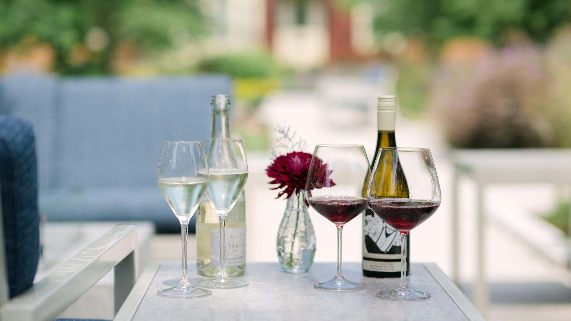 Two glasses of white wine, two glasses of red wine, and their bottles sit on an outdoor table alongside a small vase with a red flower, with blurred greenery in the background. 124 on Queen Hotel and Spa | Hotel & Spa Niagara-on-the-lake | 1 (855) 988-4552