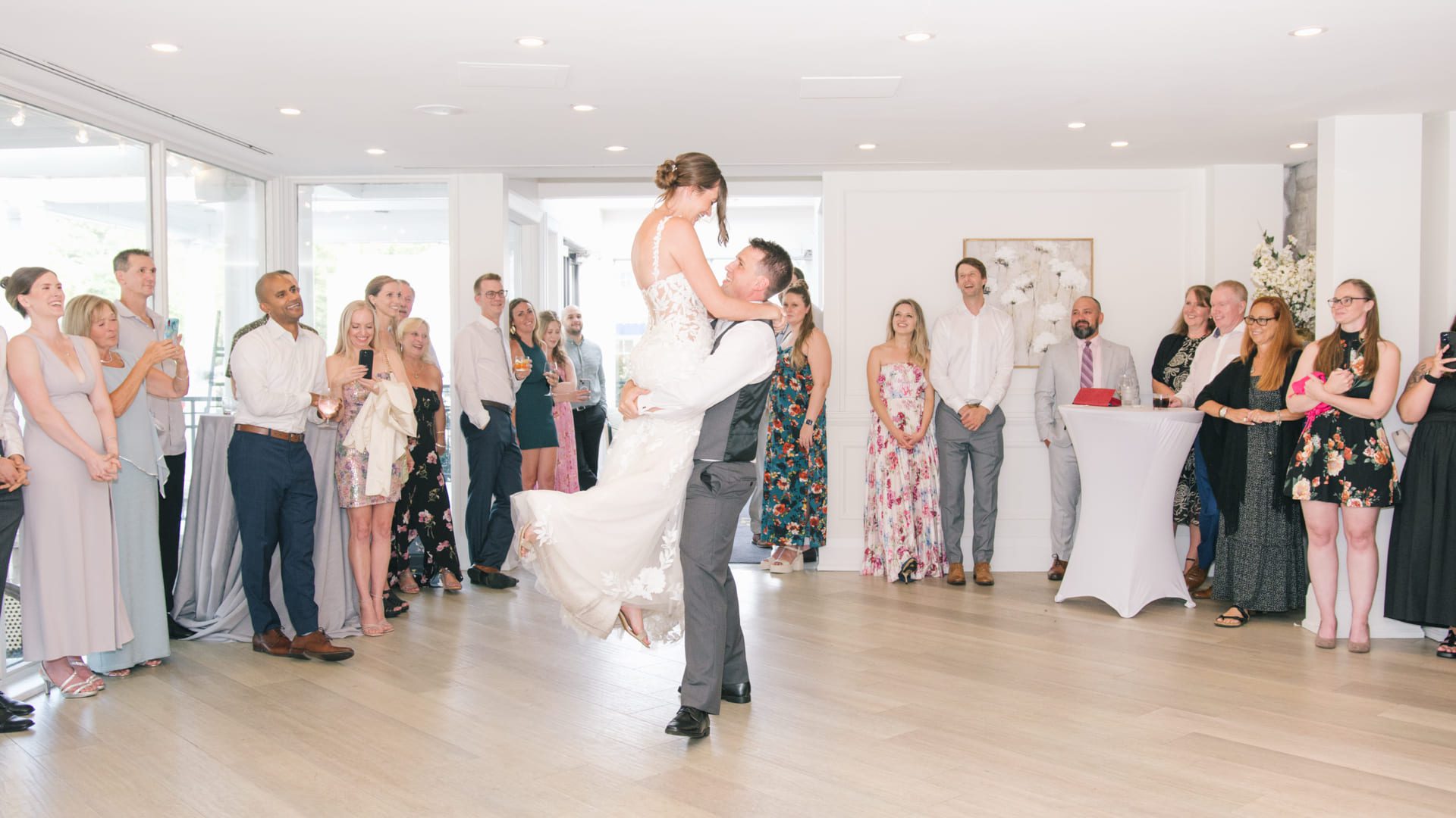 A bride and groom share their first dance as the groom lifts the bride in a bright, modern venue. Guests dressed in semi-formal attire stand in a circle around them, smiling and watching the couple. 124 on Queen Hotel and Spa | Hotel & Spa Niagara-on-the-lake | 1 (855) 988-4552