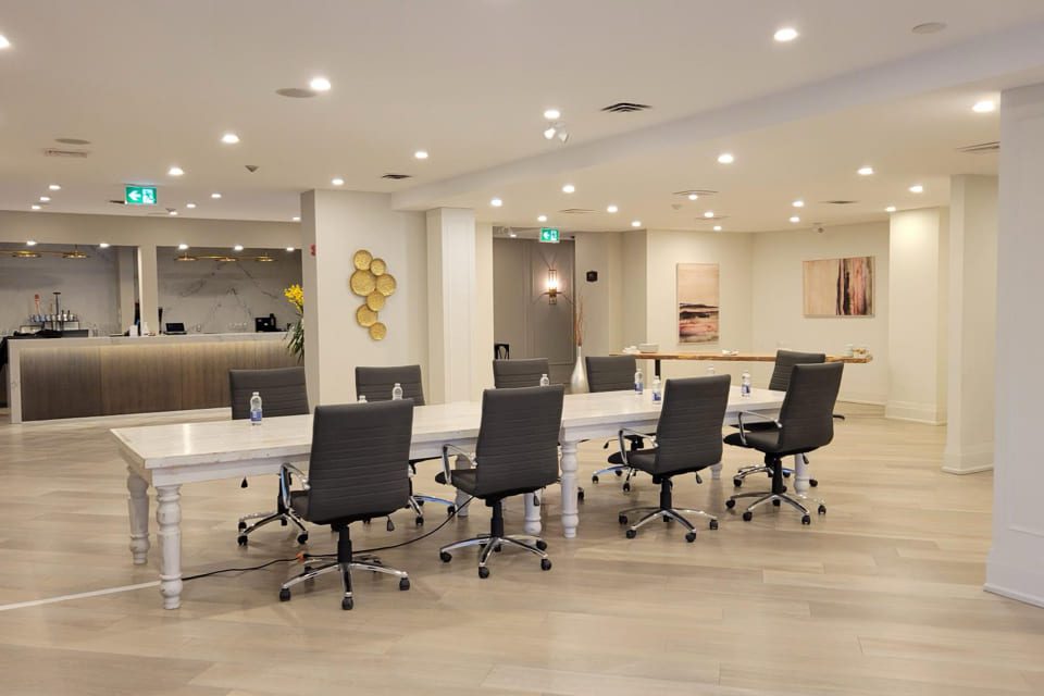 A modern conference room with a long white table, six black office chairs, bottled water on the table, abstract wall art, and a reception area with a counter in the background. The room is brightly lit with ceiling lights. 124 on Queen Hotel and Spa | Hotel & Spa Niagara-on-the-lake | 1 (855) 988-4552