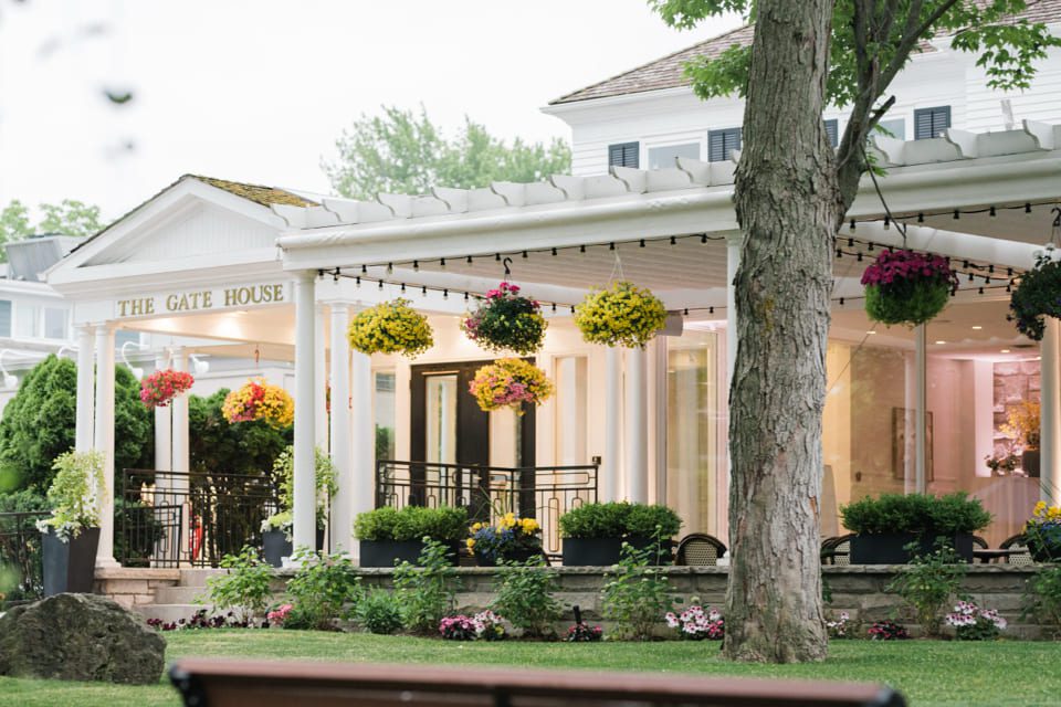 A charming white building labeled The Gate House features a covered porch adorned with hanging flower baskets, lush greenery, and blooming gardens, creating a welcoming and picturesque atmosphere. 124 on Queen Hotel and Spa | Hotel & Spa Niagara-on-the-lake | 1 (855) 988-4552