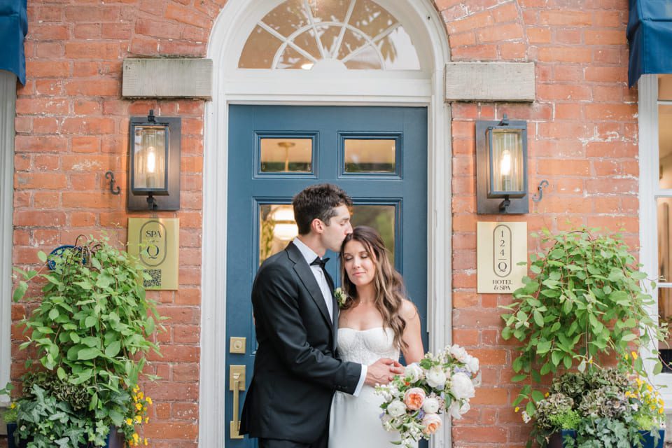 A groom in a black tuxedo kisses a bride with long hair holding a bouquet as they stand in front of a blue door framed by plants on a brick building. 124 on Queen Hotel and Spa | Hotel & Spa Niagara-on-the-lake | 1 (855) 988-4552