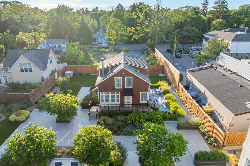 Aerial view of a two-story brown house with white trim, surrounded by a landscaped yard, patio areas, green trees, and adjacent buildings in a suburban neighborhood. 124 on Queen Hotel and Spa | Hotel & Spa Niagara-on-the-lake | 1 (855) 988-4552