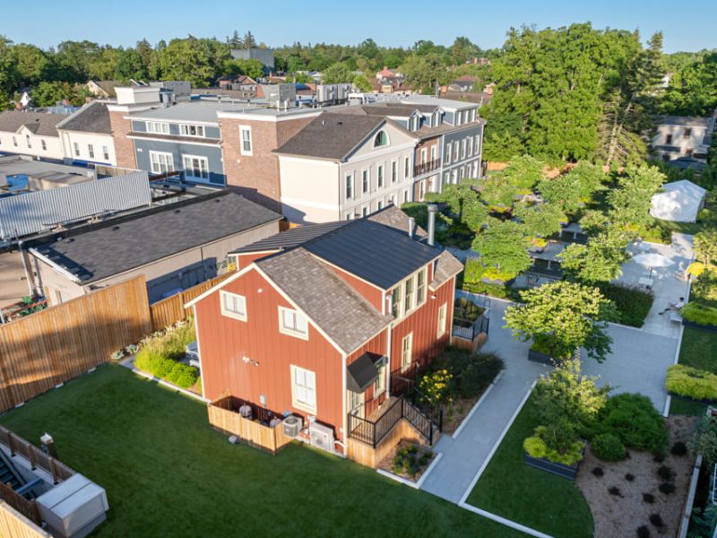 Aerial view of a red two-story house with a fenced yard, surrounded by landscaped gardens and greenery. Nearby are modern buildings and walkways with trees and outdoor seating. 124 on Queen Hotel and Spa | Hotel & Spa Niagara-on-the-lake | 1 (855) 988-4552