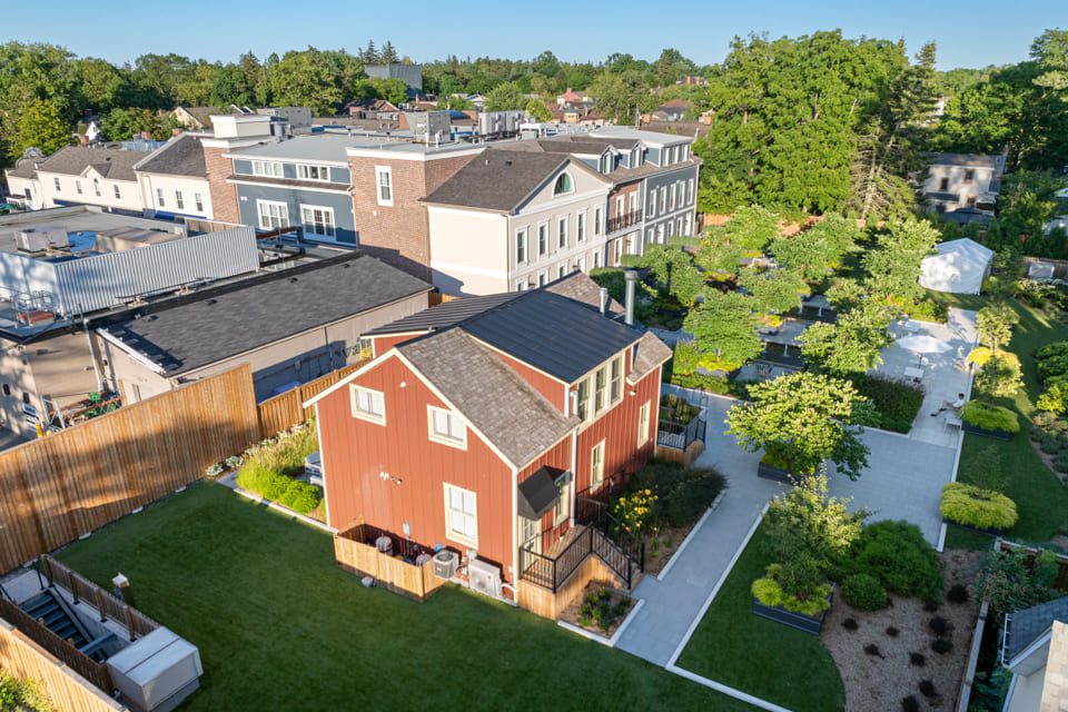 Aerial view of a red two-story house with a fenced yard, surrounded by landscaped gardens and greenery. Nearby are modern buildings and walkways with trees and outdoor seating. 124 on Queen Hotel and Spa | Hotel & Spa Niagara-on-the-lake | 1 (855) 988-4552