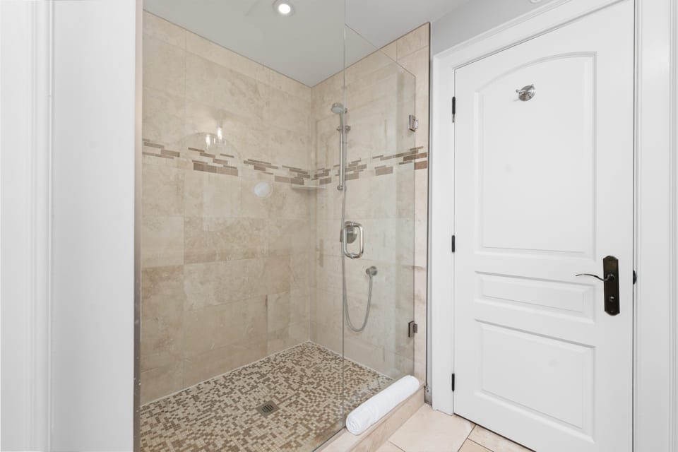 A modern bathroom with a glass-enclosed shower featuring beige tiles, a mosaic tile floor, a built-in showerhead, and a white towel on the threshold next to a white paneled door. 124 on Queen Hotel and Spa | Hotel & Spa Niagara-on-the-lake | 1 (855) 988-4552