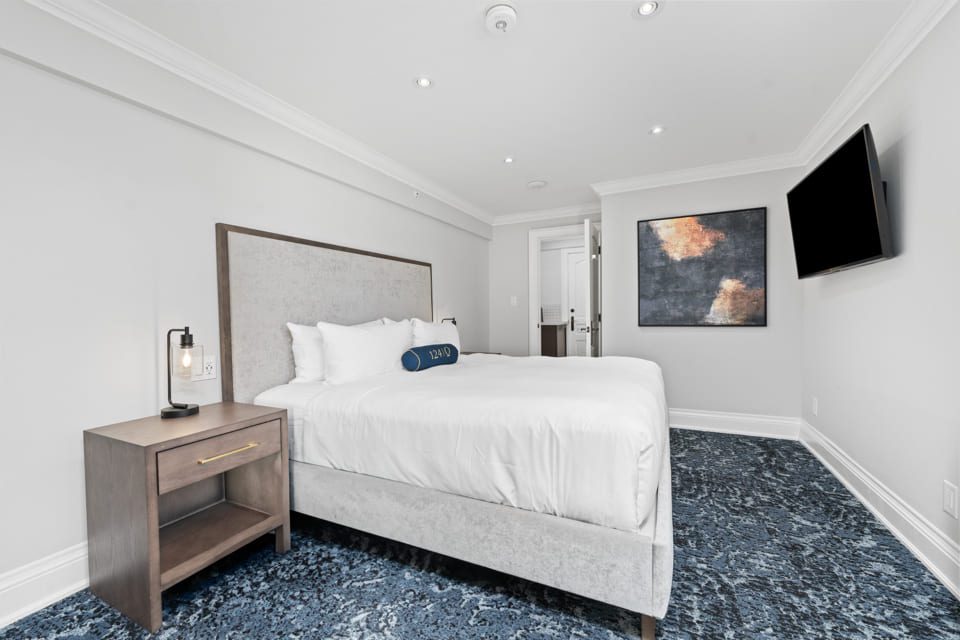 A modern bedroom with a large bed, white bedding, a gray headboard, a nightstand with a lamp, abstract wall art, and a wall-mounted TV. The room has blue patterned carpet and white walls. 124 on Queen Hotel and Spa | Hotel & Spa Niagara-on-the-lake | 1 (855) 988-4552