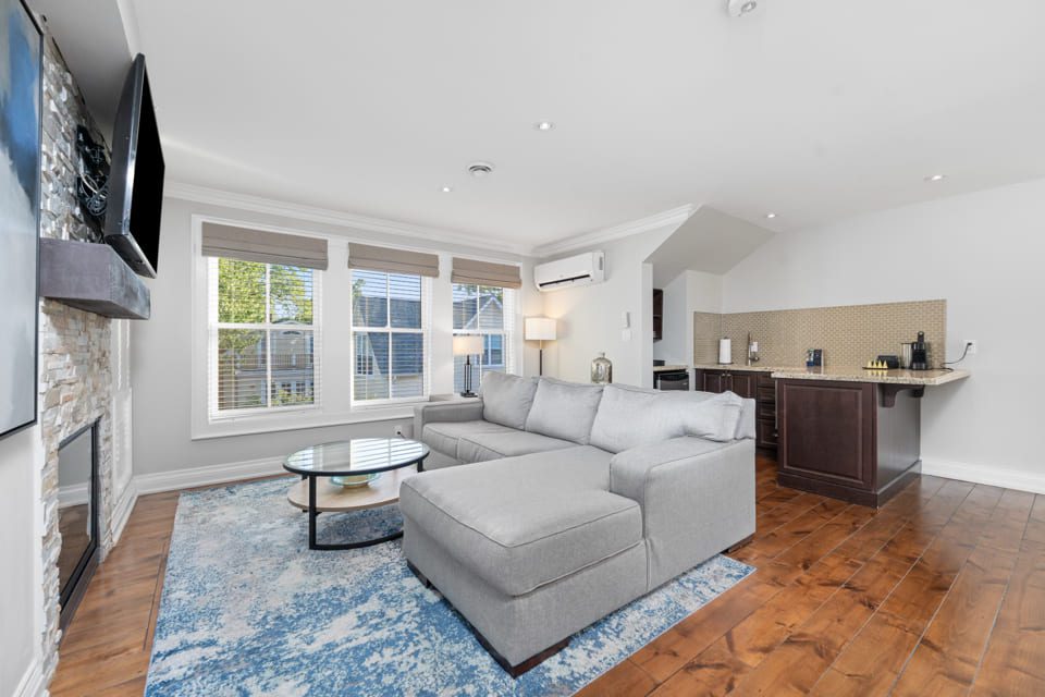 A bright living room with a large gray sectional sofa, glass coffee table, fireplace, and wall-mounted TV. There are big windows, a blue patterned rug, and a kitchenette with a breakfast bar on wooden floors. 124 on Queen Hotel and Spa | Hotel & Spa Niagara-on-the-lake | 1 (855) 988-4552