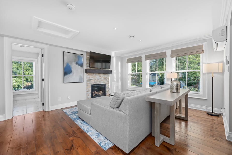 A bright, modern living room with hardwood floors, a gray sectional sofa, a fireplace with a TV above it, large windows with shades, and a console table behind the sofa. A doorway leads to another sunlit room. 124 on Queen Hotel and Spa | Hotel & Spa Niagara-on-the-lake | 1 (855) 988-4552