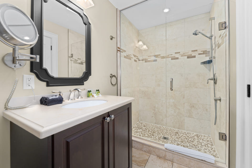 A modern bathroom with a glass-enclosed shower, beige tile walls, mosaic tile shower floor, dark wood vanity with a white countertop, wall-mounted mirror, and chrome fixtures. 124 on Queen Hotel and Spa | Hotel & Spa Niagara-on-the-lake | 1 (855) 988-4552