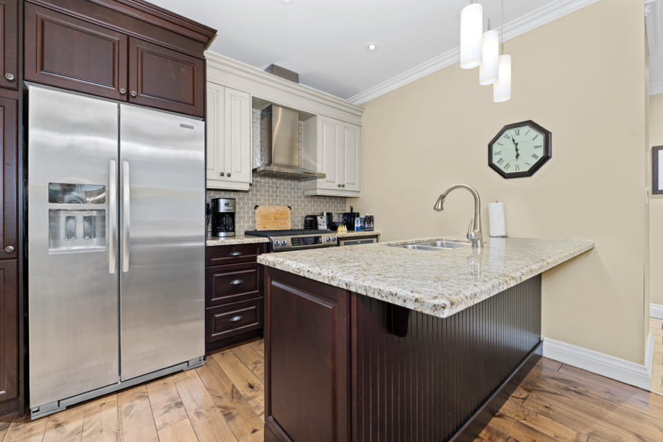 Modern kitchen with stainless steel refrigerator, stove, and range hood, white cabinets, granite island with sink, pendant lights, wooden floor, and a wall clock. 124 on Queen Hotel and Spa | Hotel & Spa Niagara-on-the-lake | 1 (855) 988-4552