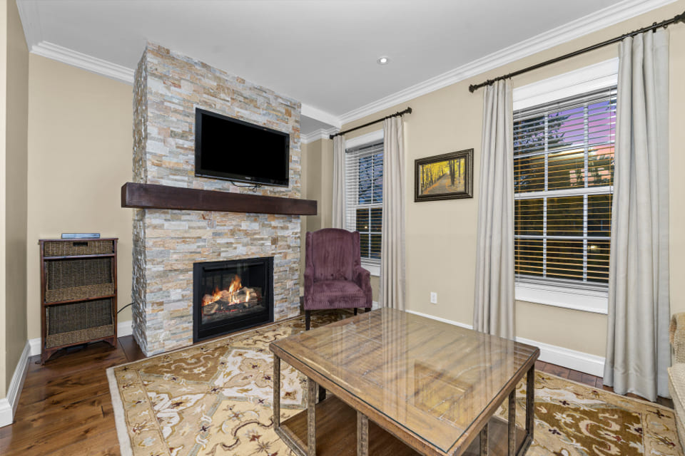 A cozy living room with a stone fireplace, a mounted TV, a purple armchair, a wooden coffee table, beige walls, two windows with curtains, and a patterned rug on a wood floor. 124 on Queen Hotel and Spa | Hotel & Spa Niagara-on-the-lake | 1 (855) 988-4552