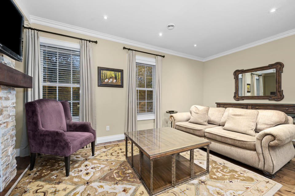 A cozy living room with a beige sofa, a purple armchair, a wooden coffee table, a patterned rug, and a large mirror above a console table. Two windows with curtains bring in natural light. 124 on Queen Hotel and Spa | Hotel & Spa Niagara-on-the-lake | 1 (855) 988-4552