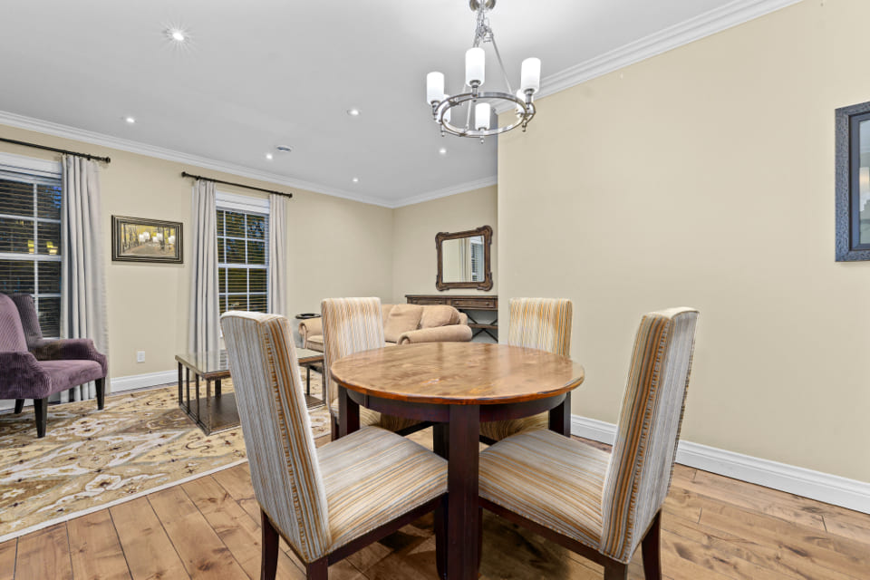 A dining area with a round wooden table and four striped upholstered chairs, adjacent to a living room with sofas, a rug, and large windows with curtains, all in a neutral color scheme. 124 on Queen Hotel and Spa | Hotel & Spa Niagara-on-the-lake | 1 (855) 988-4552