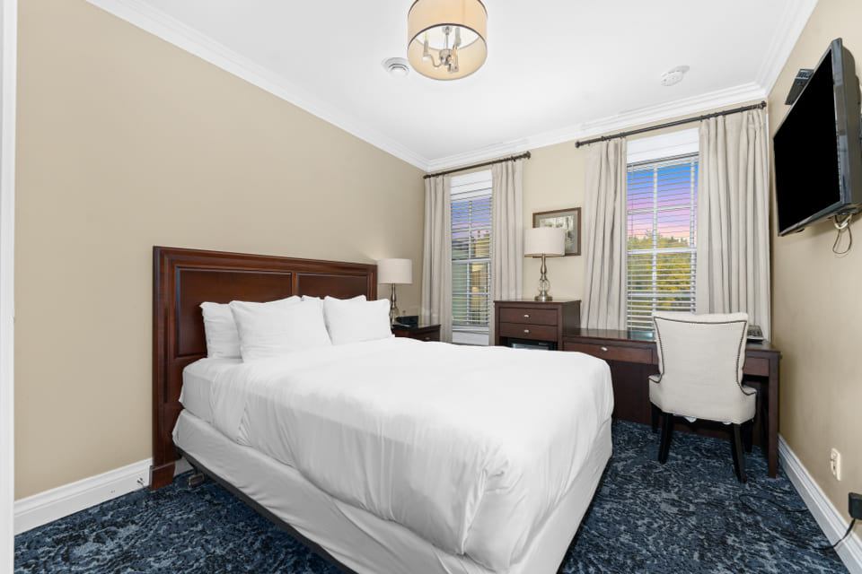 A neatly made bed with white linens in a cozy bedroom featuring beige walls, a dark wooden headboard, a desk with a chair near two tall windows, and a mounted flat-screen TV. 124 on Queen Hotel and Spa | Hotel & Spa Niagara-on-the-lake | 1 (855) 988-4552