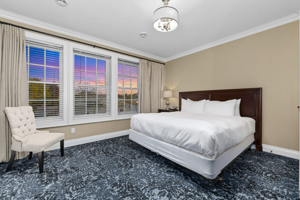 A neatly made bed with white linens sits in a clean, modern bedroom with beige walls, a patterned blue carpet, a white upholstered chair, and large windows showing a colorful sunset sky. 124 on Queen Hotel and Spa | Hotel & Spa Niagara-on-the-lake | 1 (855) 988-4552