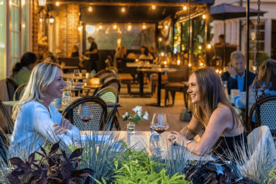 Two women sit at an outdoor restaurant table, smiling and talking. They have wine glasses and a small flower vase on the table. Other diners and warm string lights create a cozy evening atmosphere in the background. 124 on Queen Hotel and Spa | Hotel & Spa Niagara-on-the-lake | 1 (855) 988-4552