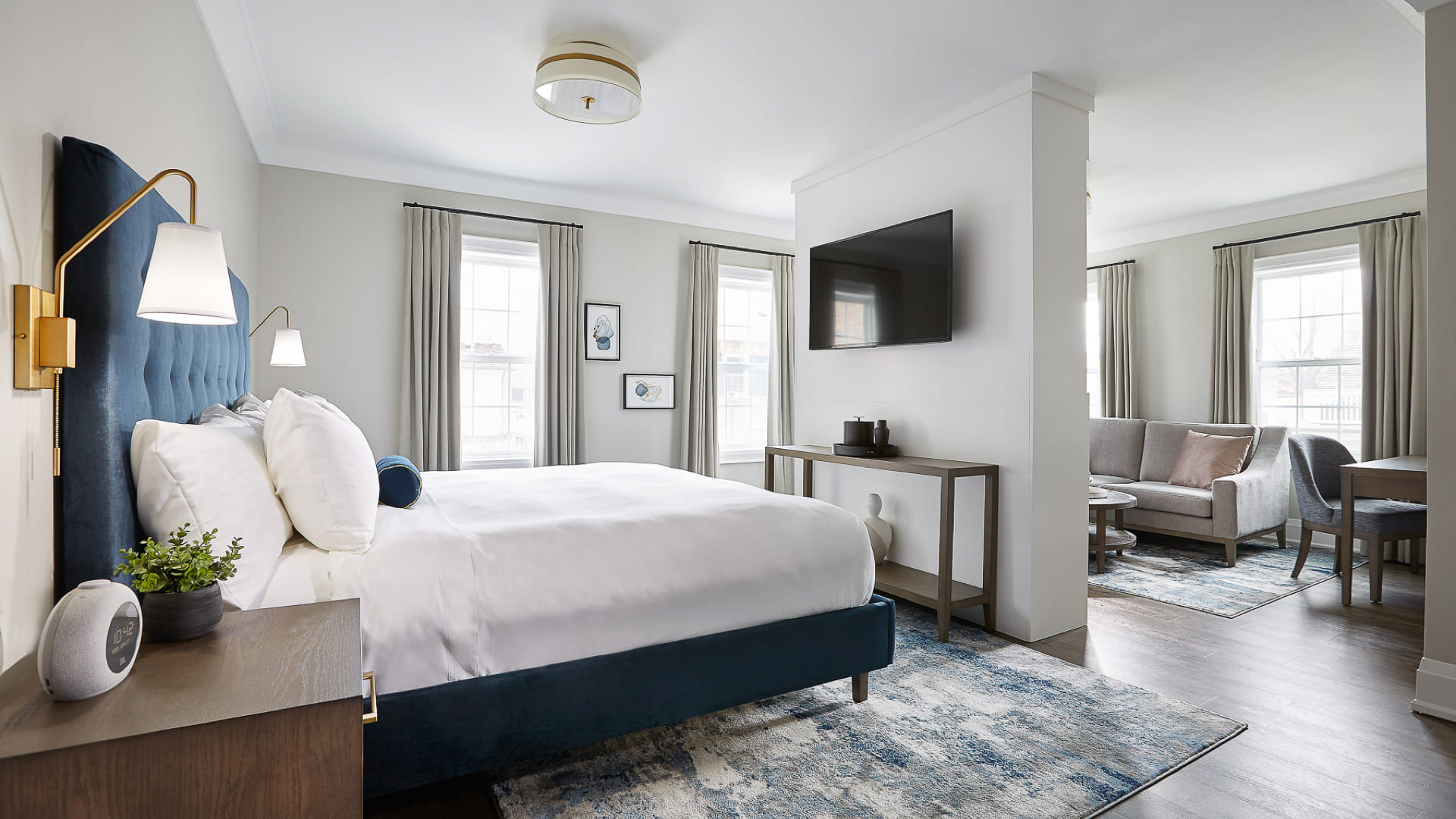 Bright, modern hotel room with a large bed, blue velvet headboard, bedside tables, and wall-mounted TV. Enjoy a cozy sitting area and large windows. Discover comfort and style with exclusive Niagara-on-the-Lake hotel deals. 124 on Queen Hotel and Spa | Hotel & Spa Niagara-on-the-lake | 1 (855) 988-4552