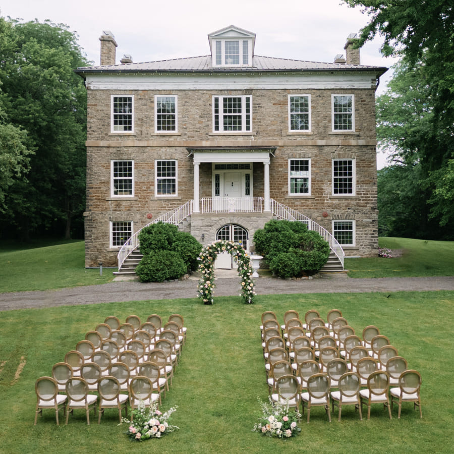 Outdoor wedding ceremony setup on a lawn in front of a large, historic stone building, with rows of chairs facing a floral arch at the base of a staircase leading to the entrance. Lush greenery surrounds the scene. 124 on Queen Hotel and Spa | Hotel & Spa Niagara-on-the-lake | 1 (855) 988-4552