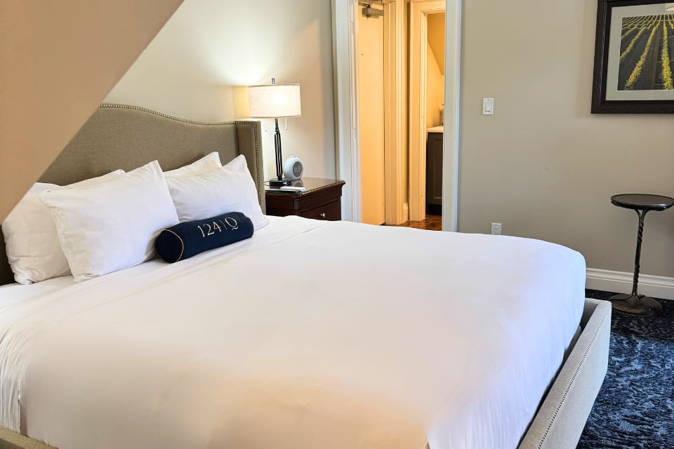 A neatly made bed with white linens and a blue decorative pillow sits next to a nightstand with a lamp in a cozy, well-lit bedroom. An open door reveals a glimpse of a bathroom. 124 on Queen Hotel and Spa | Hotel & Spa Niagara-on-the-lake | 1 (855) 988-4552