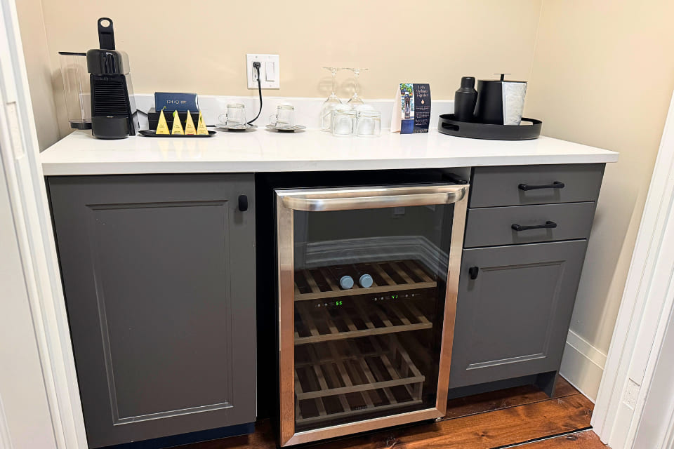 A modern kitchenette with a coffee maker, cups, glasses, condiments, and a tray on a white countertop. Below are grey cabinets and a glass-front mini fridge with wooden shelves. 124 on Queen Hotel and Spa | Hotel & Spa Niagara-on-the-lake | 1 (855) 988-4552