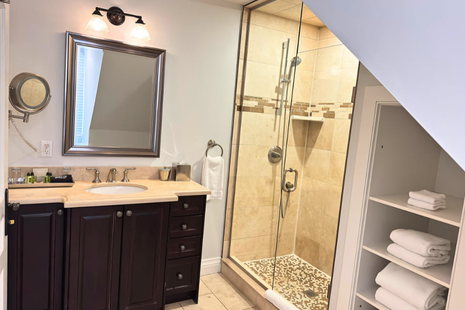 A modern bathroom with a dark wood vanity, beige countertop, large mirror, glass-enclosed tiled shower, and open white shelves stacked with folded white towels. 124 on Queen Hotel and Spa | Hotel & Spa Niagara-on-the-lake | 1 (855) 988-4552