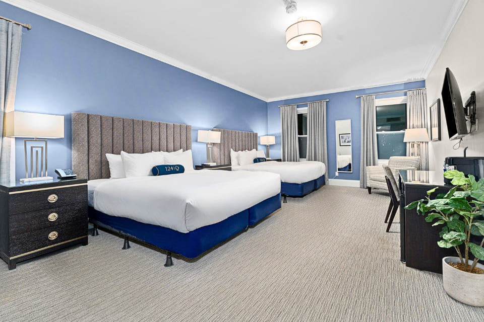 A spacious hotel room with two large beds, blue accent wall, nightstands with lamps, a wall-mounted TV, a desk and chair, large windows with gray curtains, and a potted plant in the corner. 124 on Queen Hotel and Spa | Hotel & Spa Niagara-on-the-lake | 1 (855) 988-4552