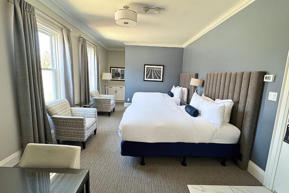 Hotel room with two double beds, gray upholstered headboards, white bedding, a nightstand with a lamp, two patterned armchairs, gray carpet, and framed photos on pale gray walls. Large windows bring in natural light. 124 on Queen Hotel and Spa | Hotel & Spa Niagara-on-the-lake | 1 (855) 988-4552